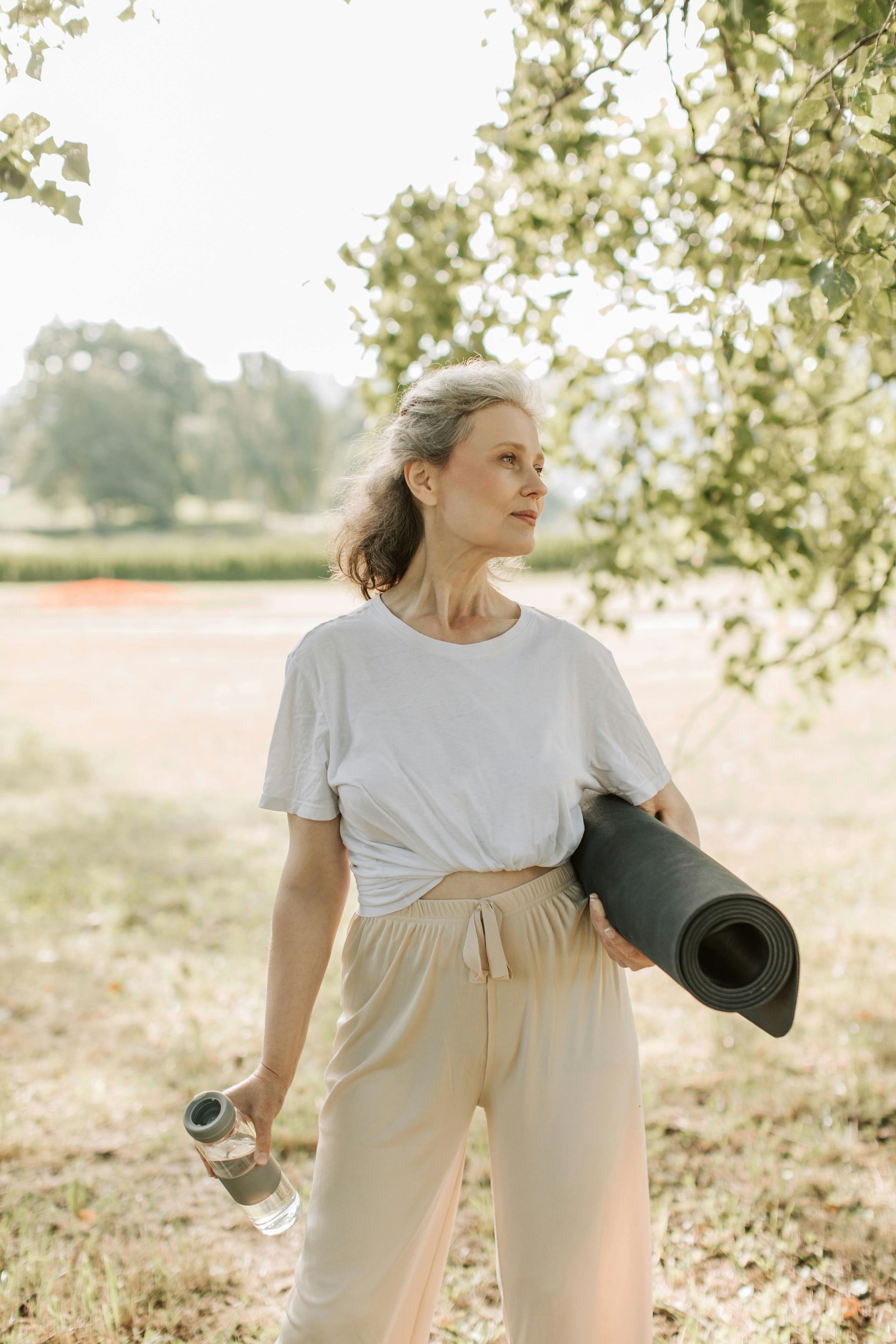 A woman is holding a yoga mat and a water bottle in a park.