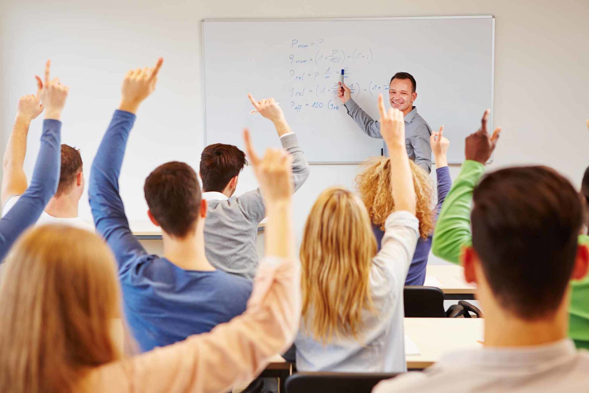 students lifting hands in college class with teacher on whiteboard