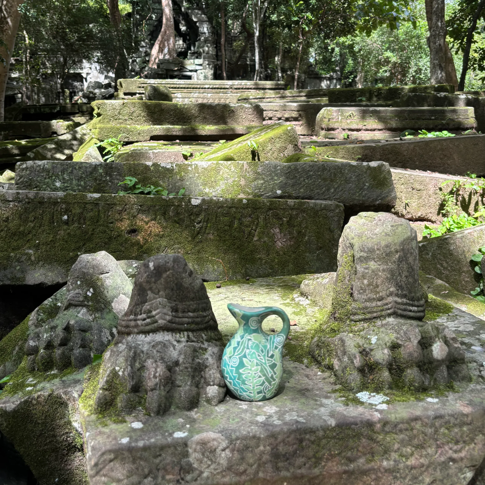Ancient stone structures, a turquoise vase, and two stone figures amid a lush forest in Cambodia.