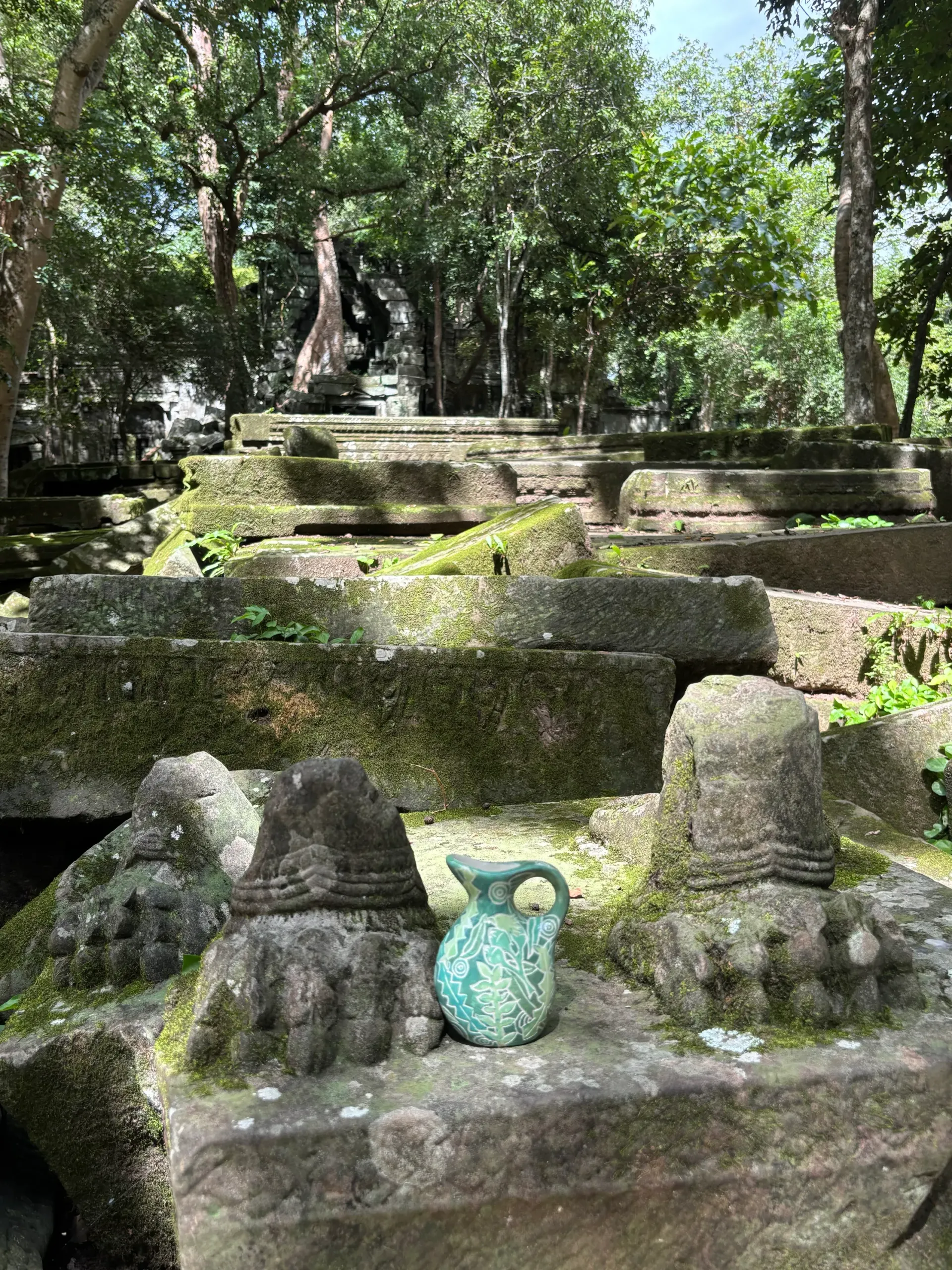 Ancient stone ruins overgrown with moss, a turquoise pitcher sits on a weathered block, green trees in the background.