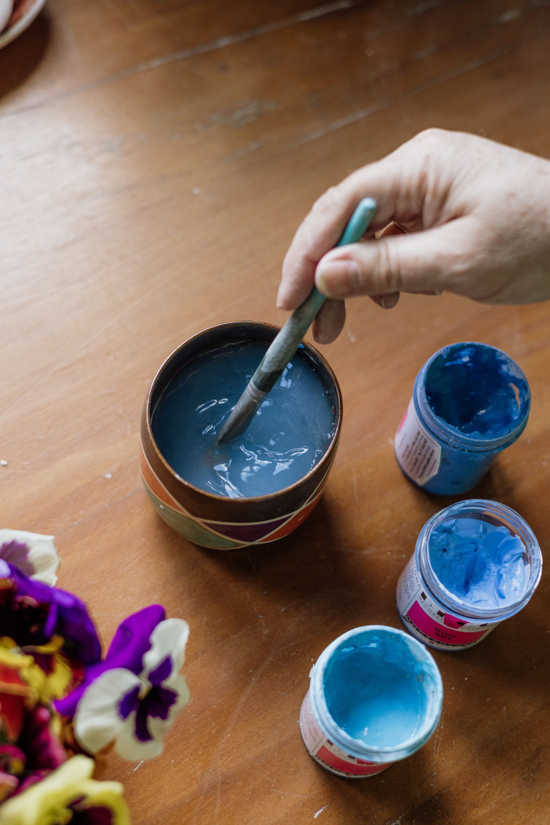 Paint brush being washed in a cup next to tubs of blue coloured underglaze and some flowers.