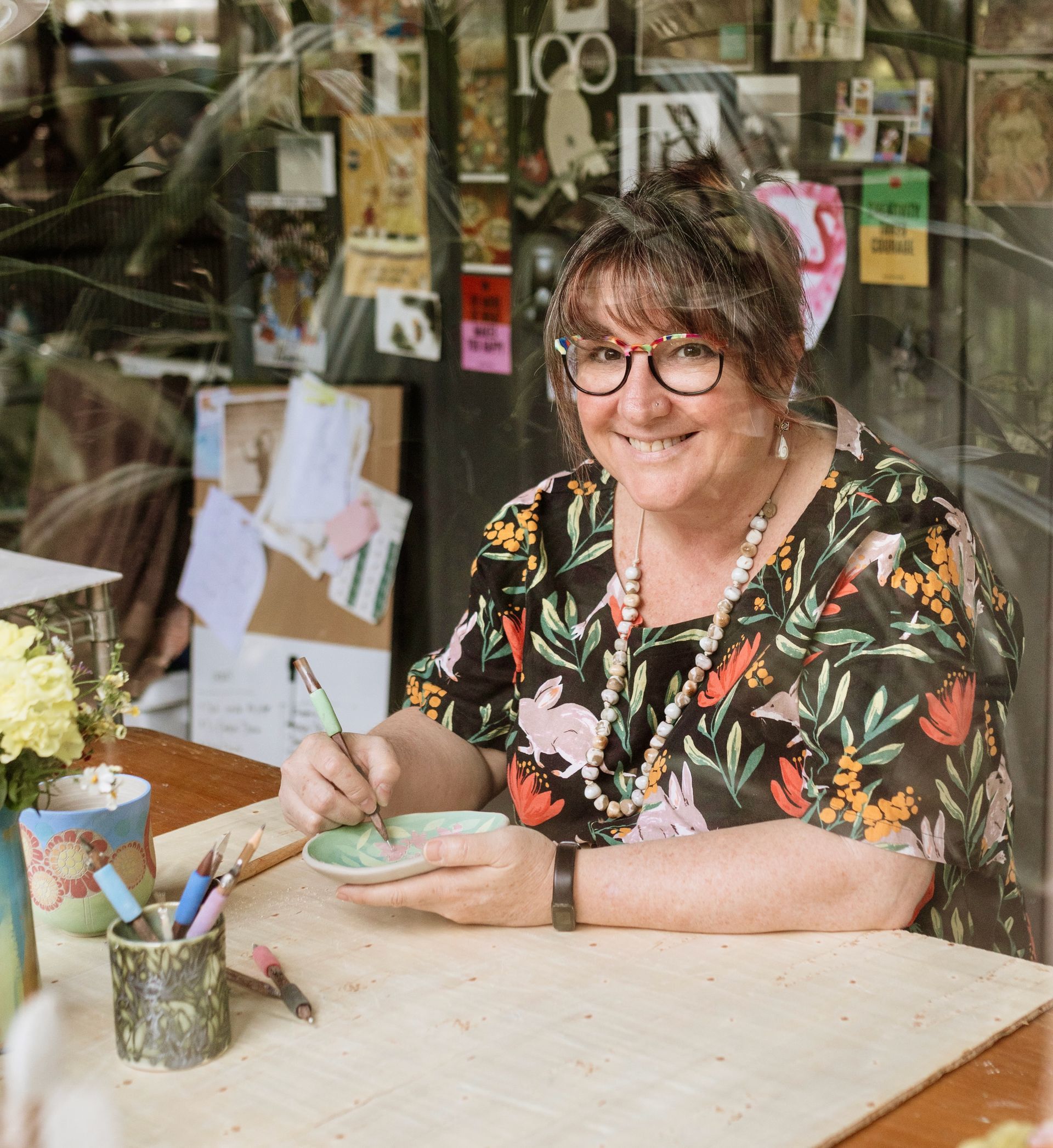 Rhianon sitting at a workbench making ceramics and smiling at the camera
