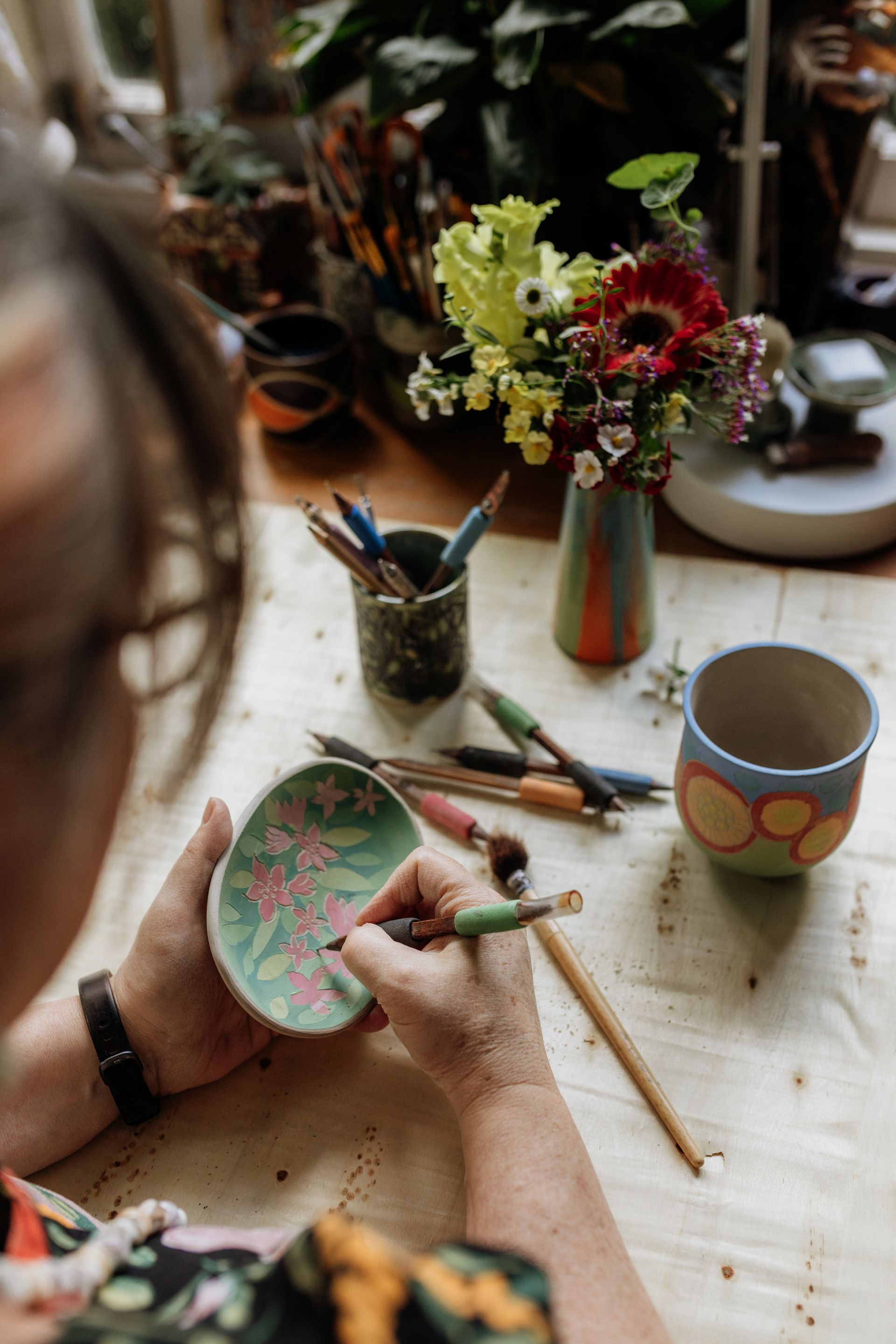 Hands carving a design on a floral dish with tools and other cups in the background