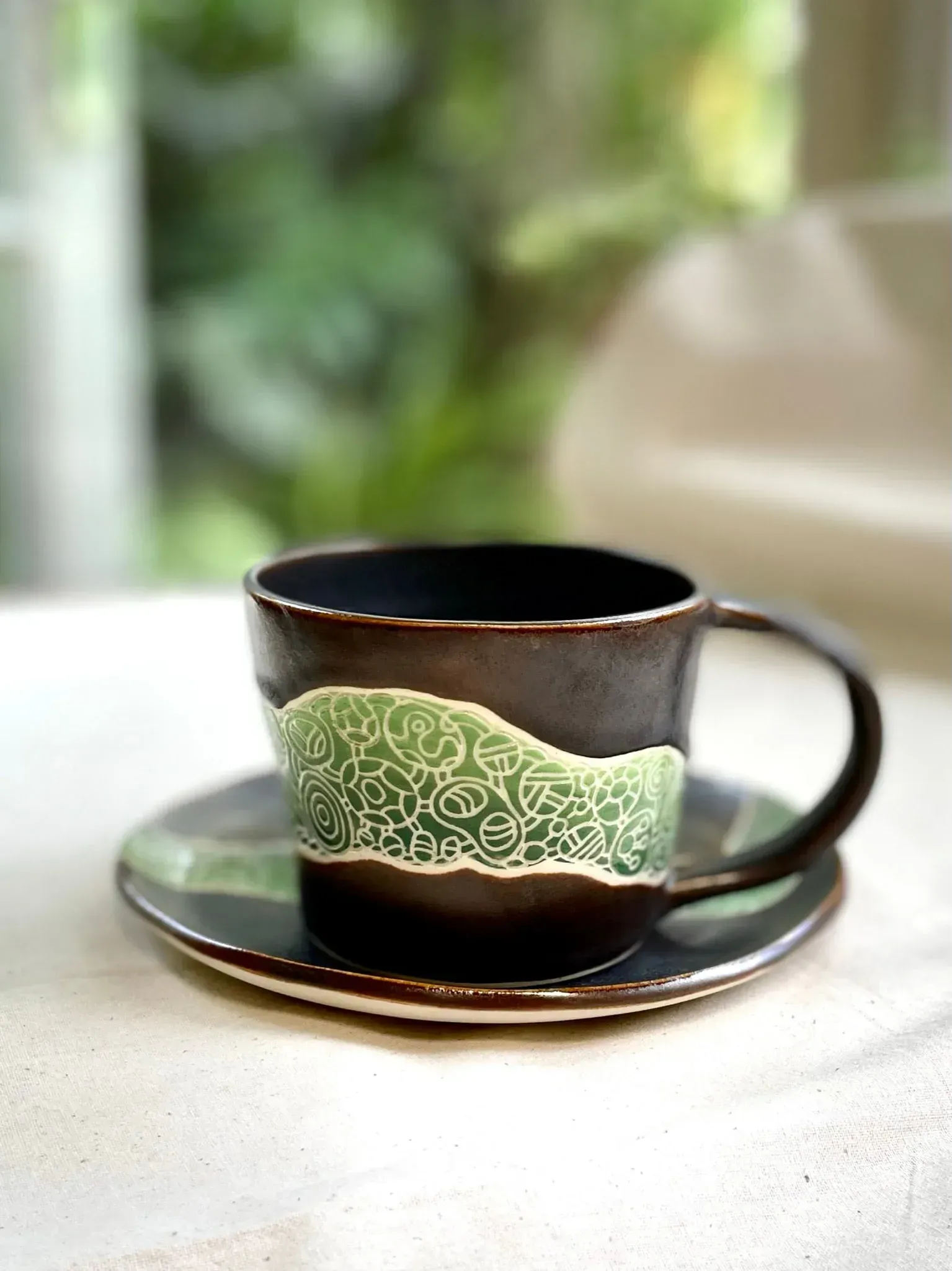 Coffee cup and saucer, dark brown with green textured band, set on a white surface, blurred green background.