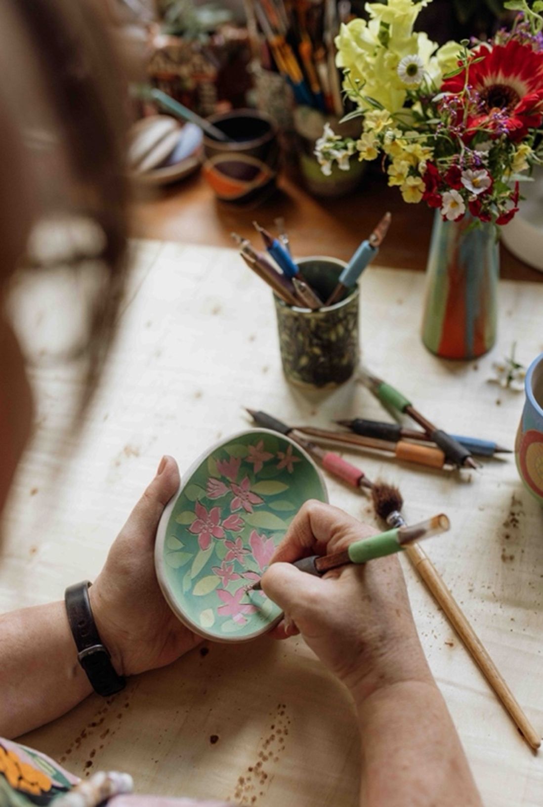 A person paints a ceramic bowl with flowers, next to vases and art tools on a wooden table.