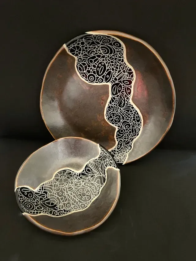 Two plates with an intricately carved black band surrounded by rich brown glaze. The carved band has a pattern of lots of small bubble shapes each with a unique carved pattern, and each connected to all of the bubbles around them.