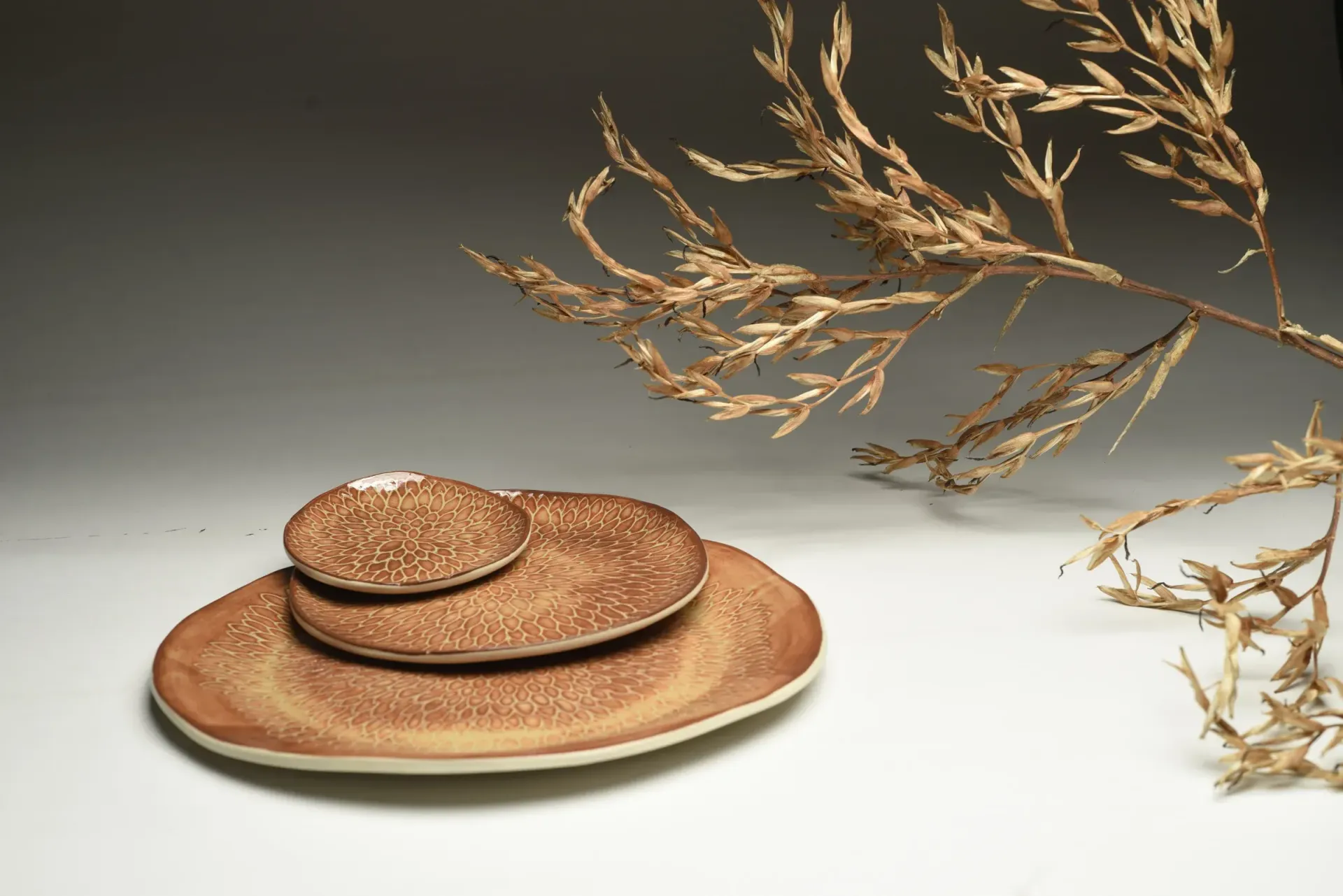 Three creamy brown ceramic plates carved with lotus leaf texture, stacked, next to dry, beige grass sprigs on a white and gray background.