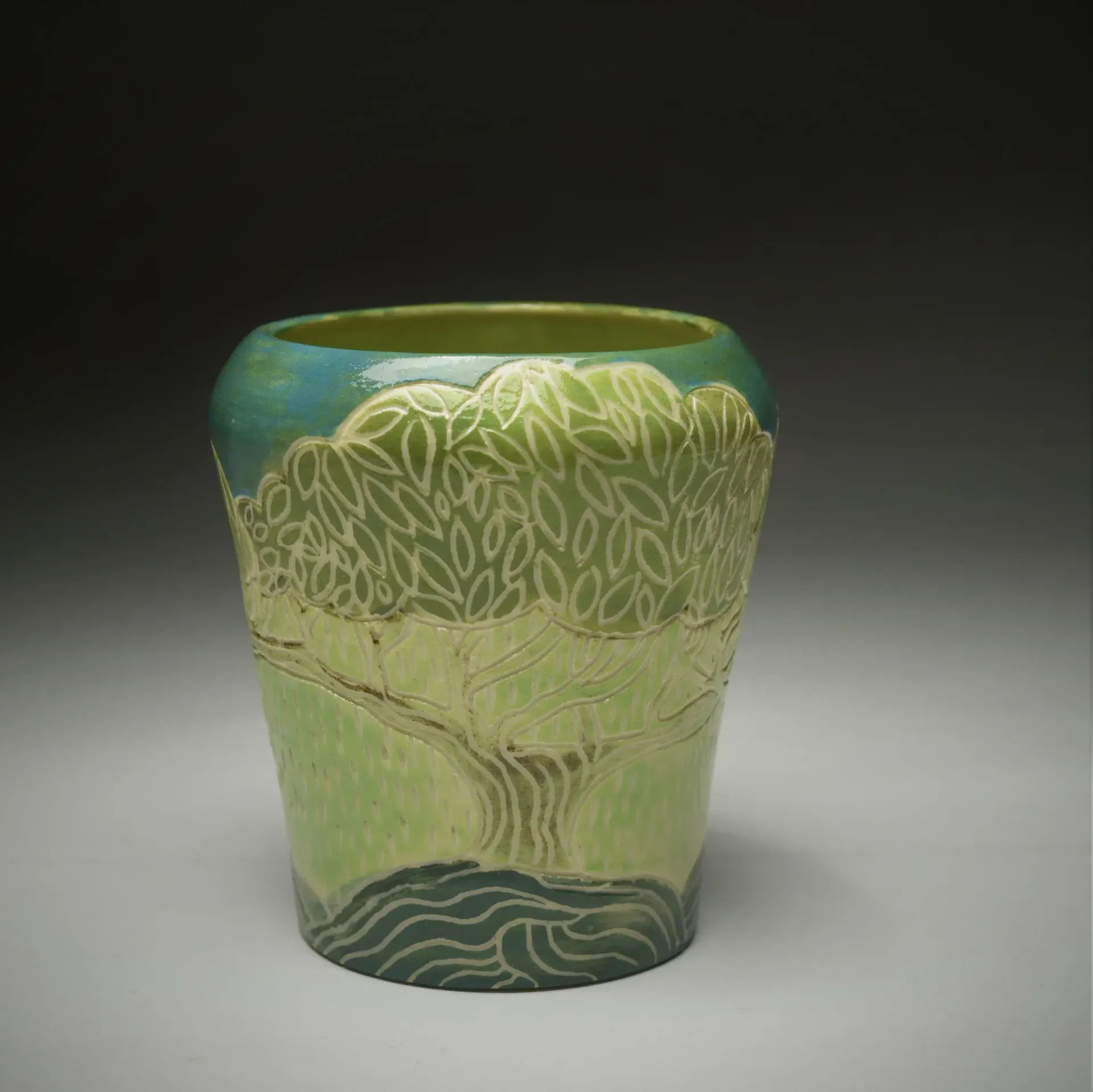 Ceramic vase, etched with trees that have interconnected limbs. Green and blue hues, tree and water details.