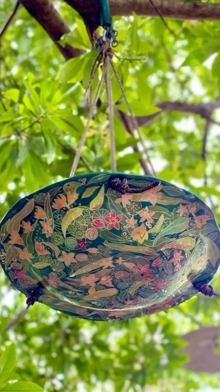 Bird feeder, oval-shaped, hanging from a tree branch, decorated with floral patterns in green, red, and yellow.