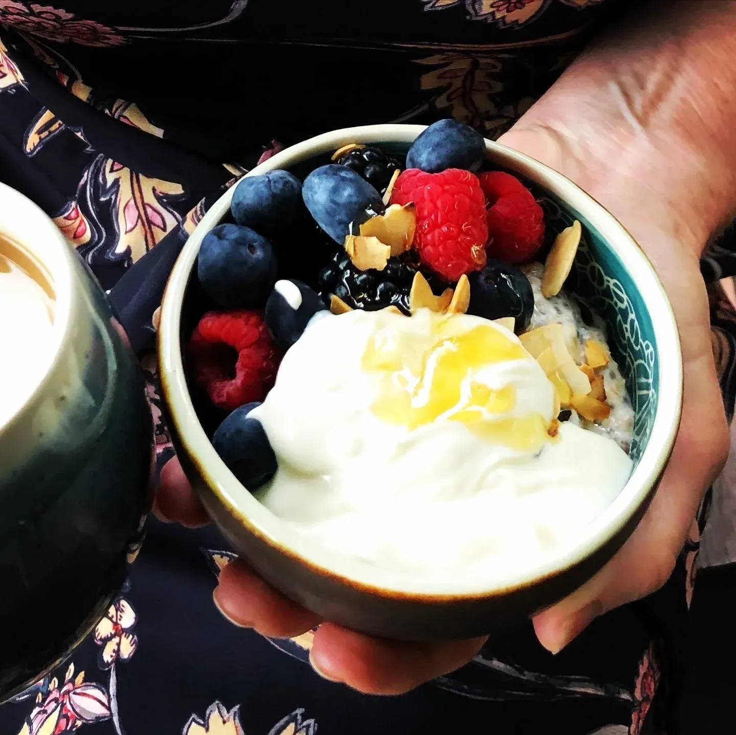 Person holding a bowl of yogurt topped with berries and a drizzle of honey; coffee cup to the left.