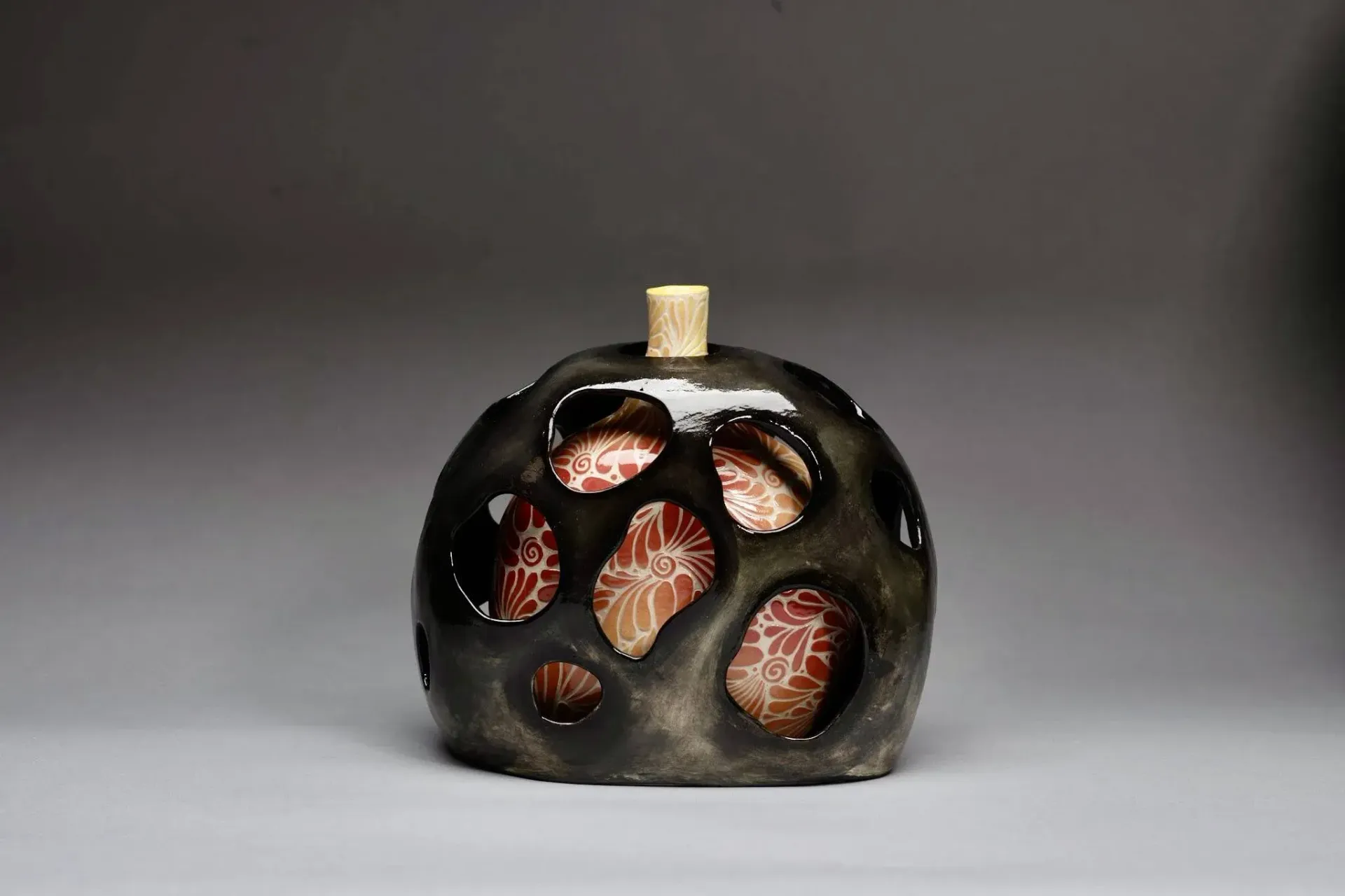Ceramic vessel with dark exterior and irregularly shaped holes; beige interior visible.