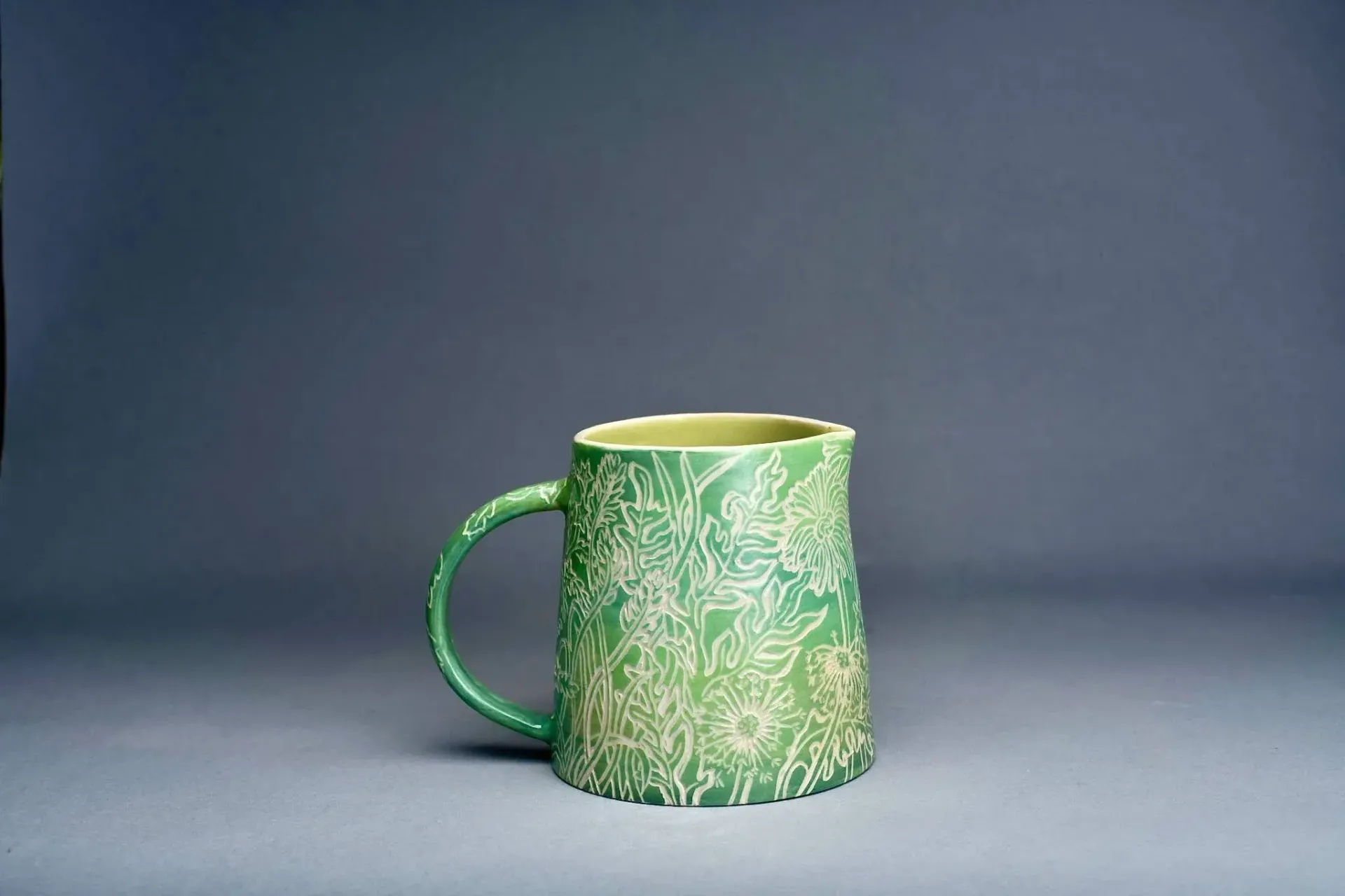 Green ceramic mug with intricate white floral design against a gray backdrop.