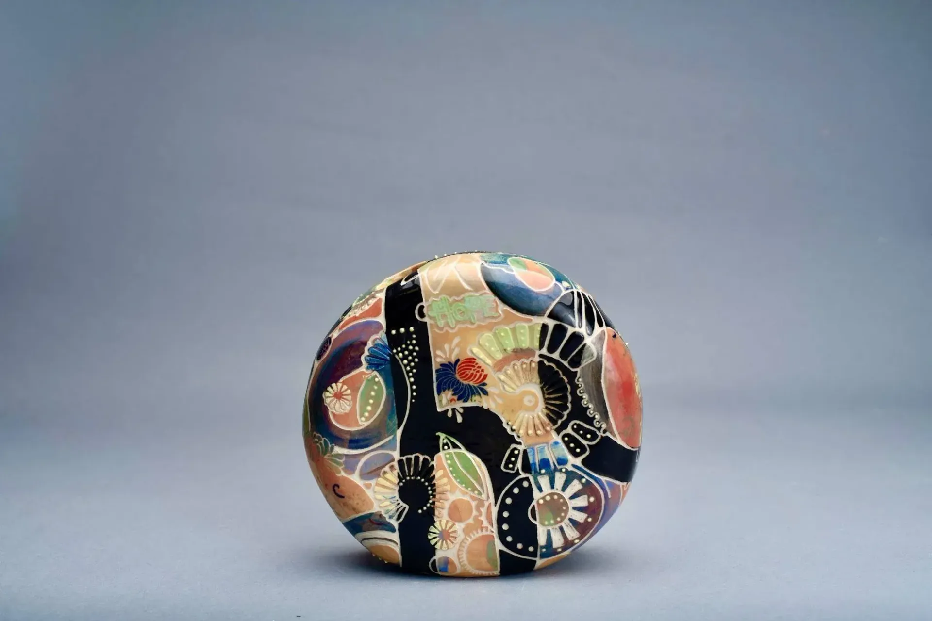 Decorative, round, patterned box with a gold base, and black, blue, and red designs against a cream background.