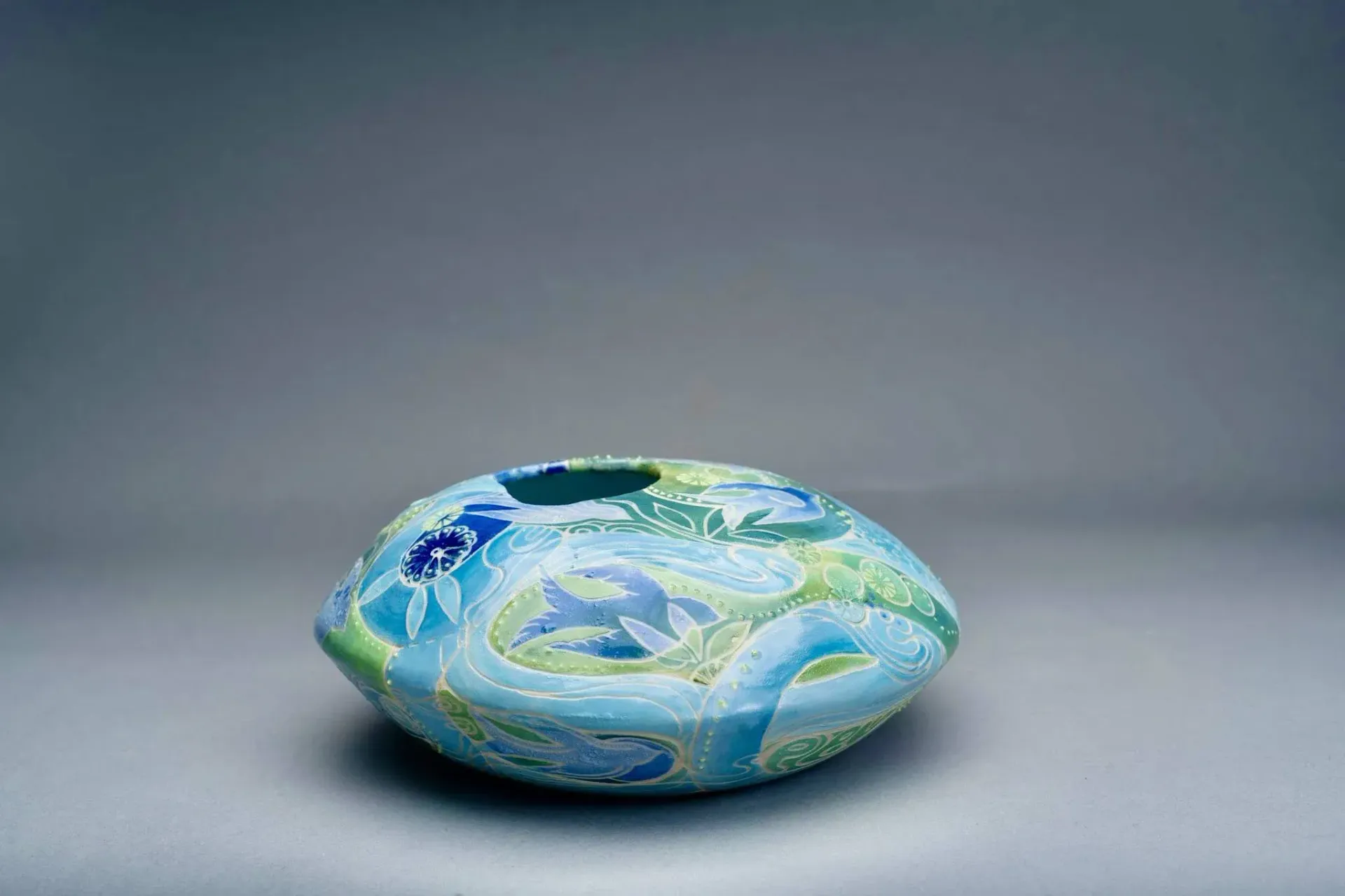 Blue and green oval glass vase with floral patterns, set against a gray background.