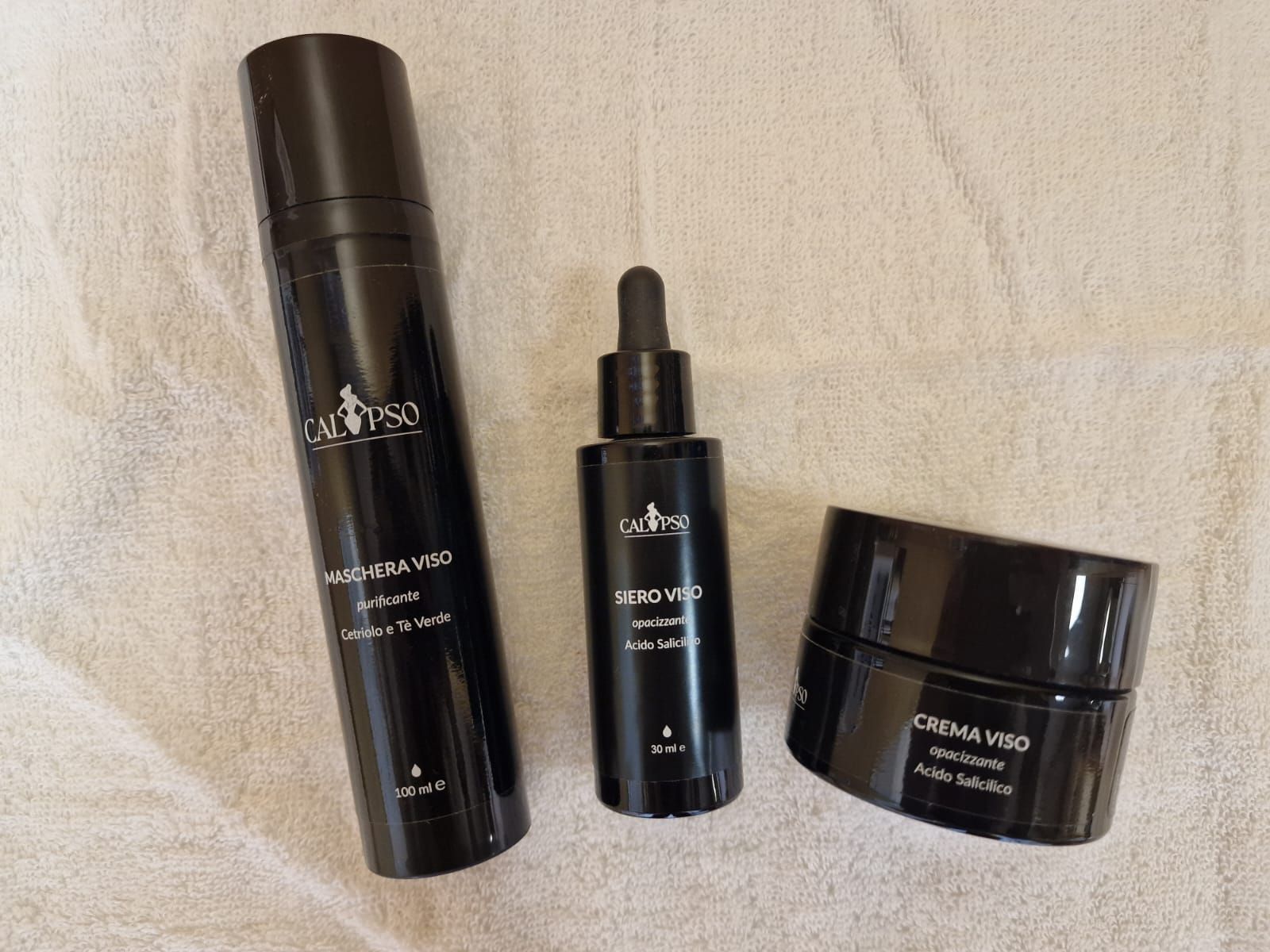 Tre bottiglie nere di cosmetici sono appoggiate su un asciugamano bianco.
