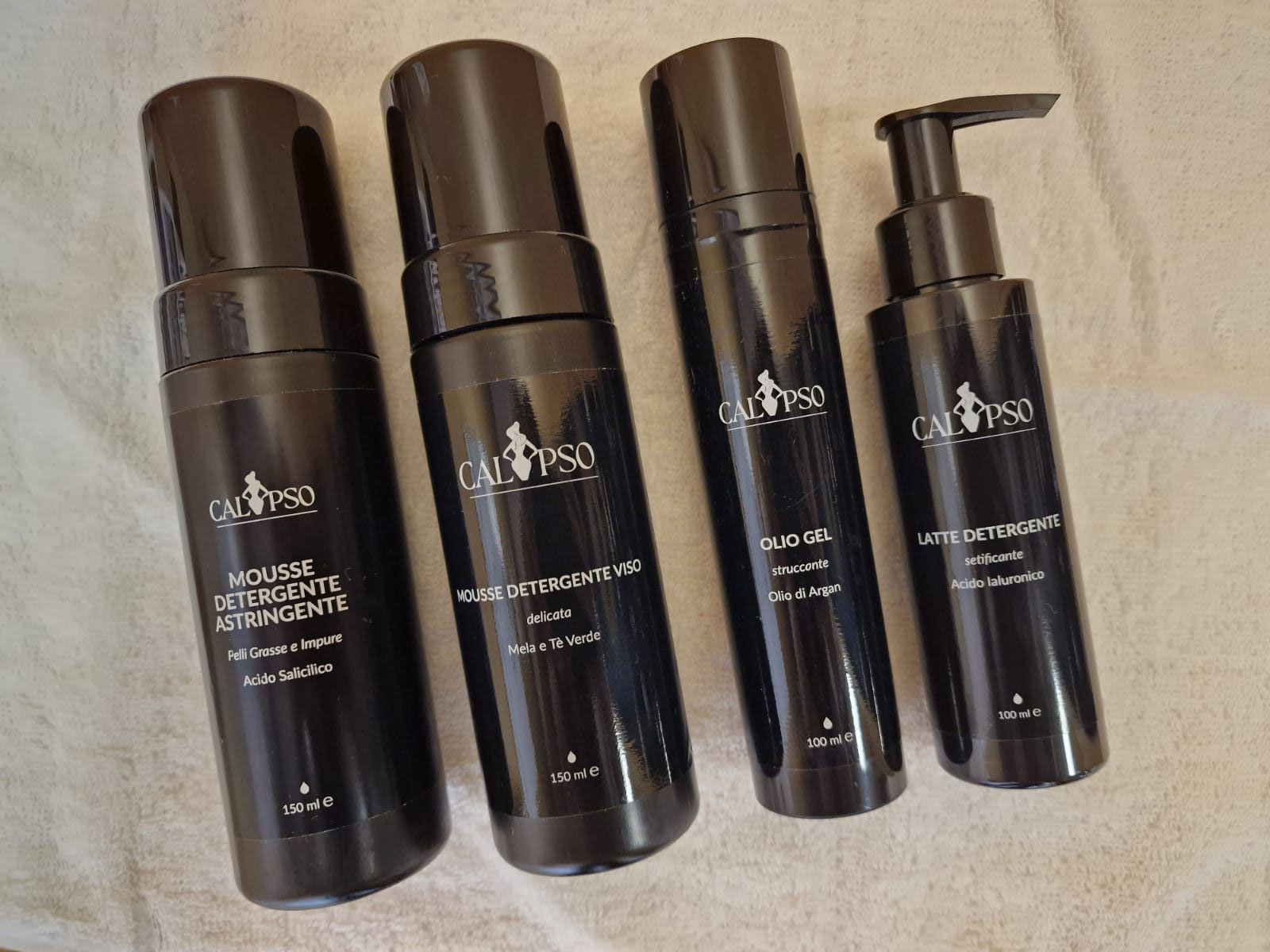 Quattro bottiglie nere di cosmetici sono appoggiate su un asciugamano bianco.