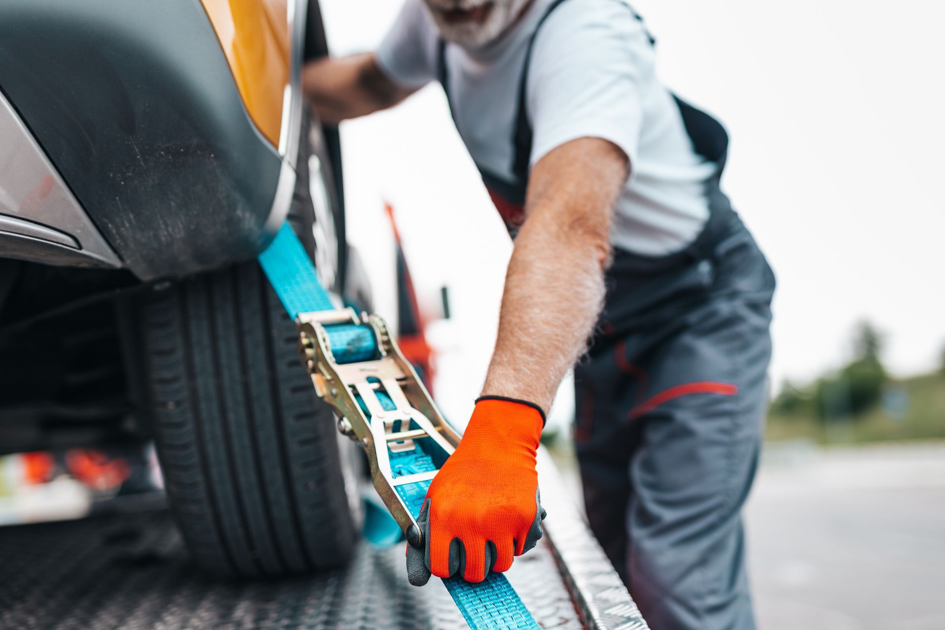 Man securing a car wheel with a blue strap on a tow truck, wearing orange gloves.