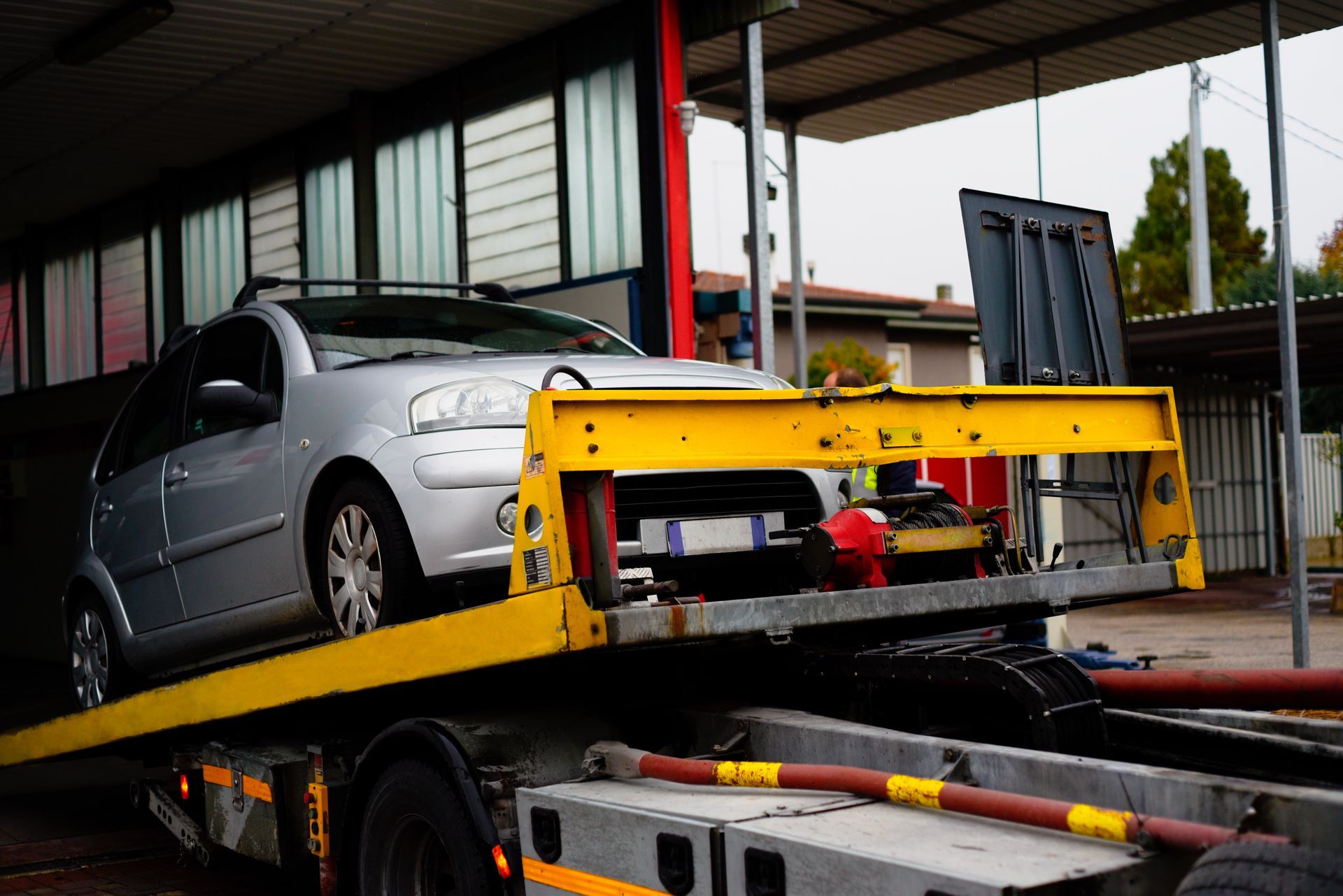 Silver car loaded on a yellow tow truck bed in front of a garage.