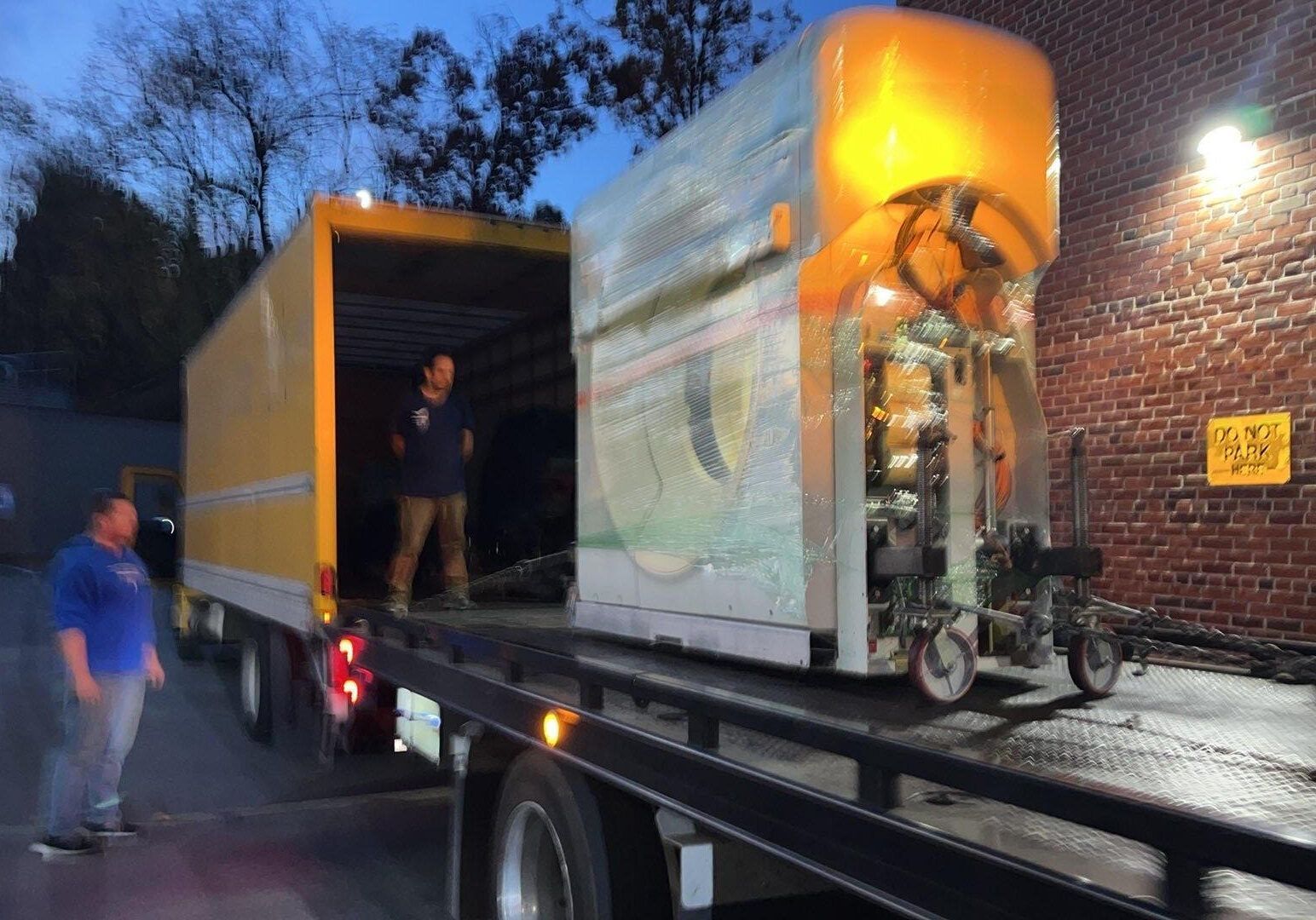 Two men loading a lawnmower into a trailer outdoors. One is guiding it, the other is assisting.