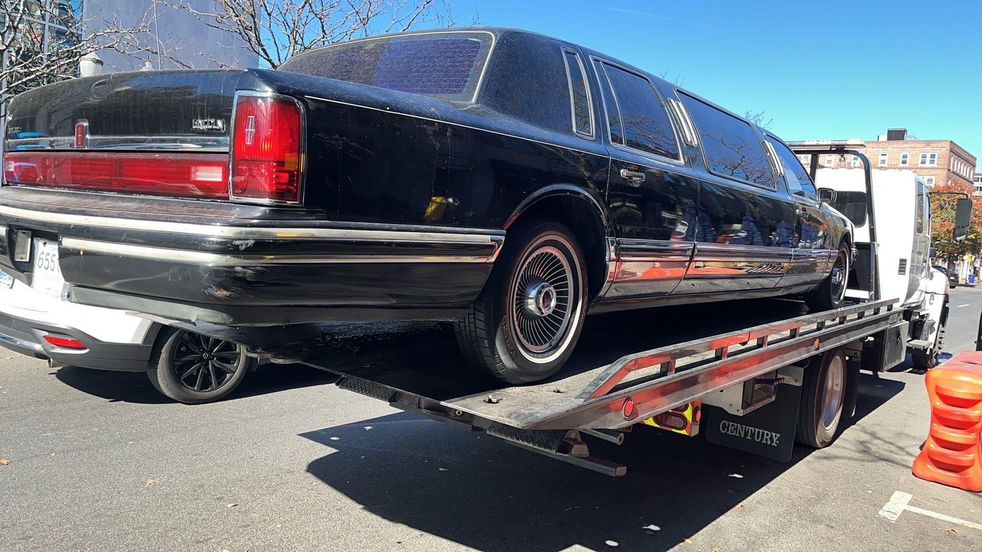 Tow truck operator loading a white car onto the flatbed; forest background.