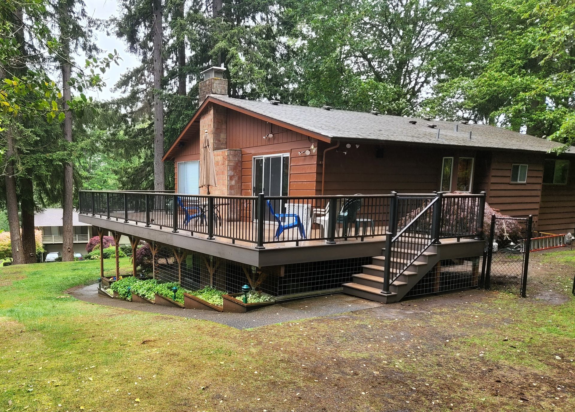 Brown house with a large deck and stairs. Green lawn with trees.