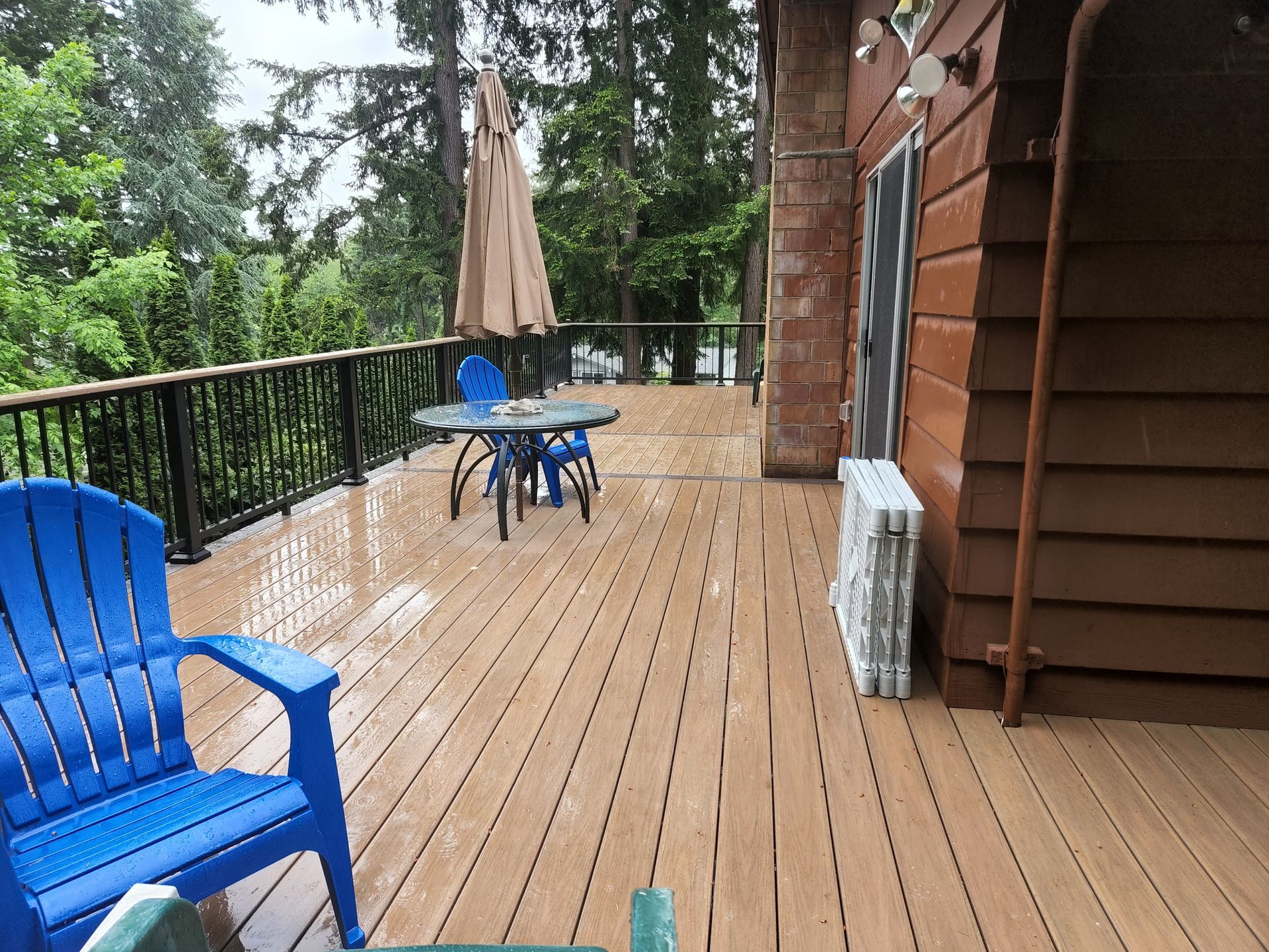A wooden deck with blue chairs, table, and umbrella in the rain, overlooking a forested area.