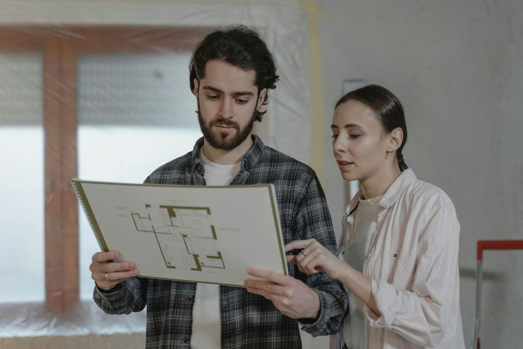 Couple reviewing a blueprint, pointing, discussing home renovation in a room covered in plastic.