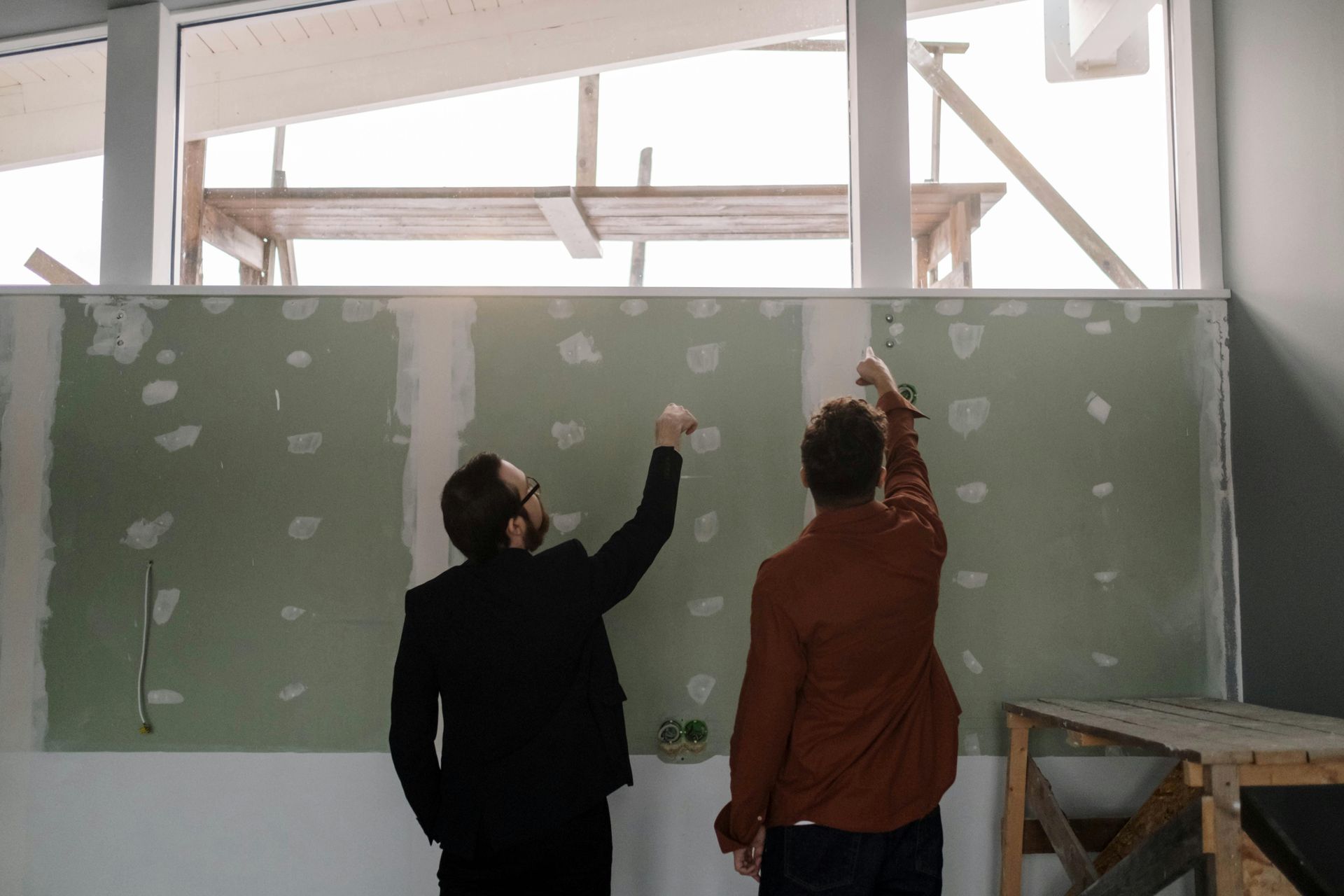 Two men pointing at a green wall, under a large window, in a room under construction.