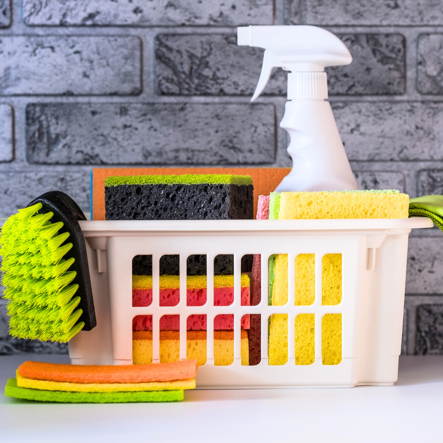 Materiais de limpeza em uma cesta branca: borrifador, esponjas, escova contra uma parede de tijolos.