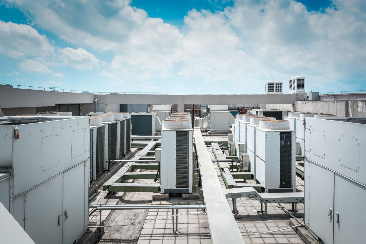 Rooftop view of commercial air conditioning units, pipes, and walkways under a partly cloudy sky.