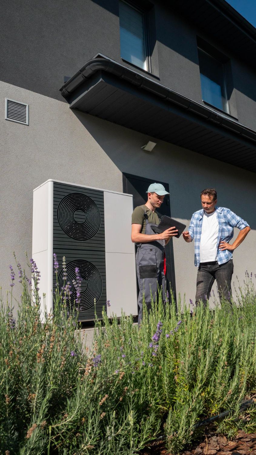 Two men near a heat pump unit installed on a house, one holding a tablet.