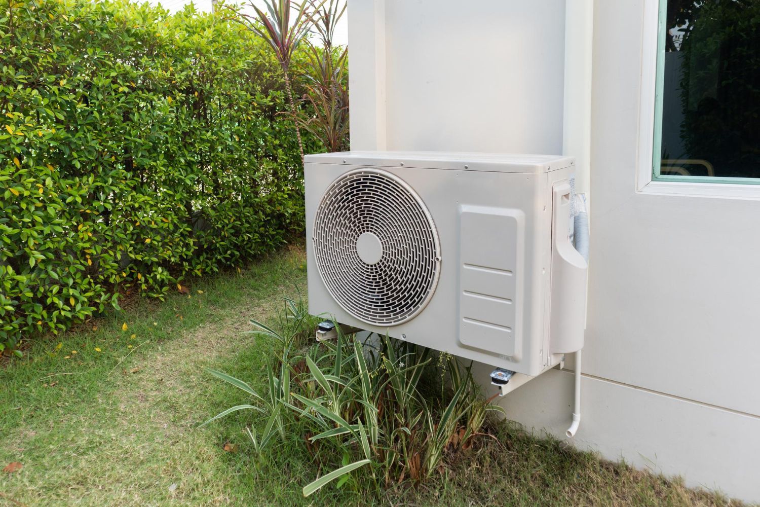 Outdoor air conditioning unit mounted on a white building wall next to a bush.