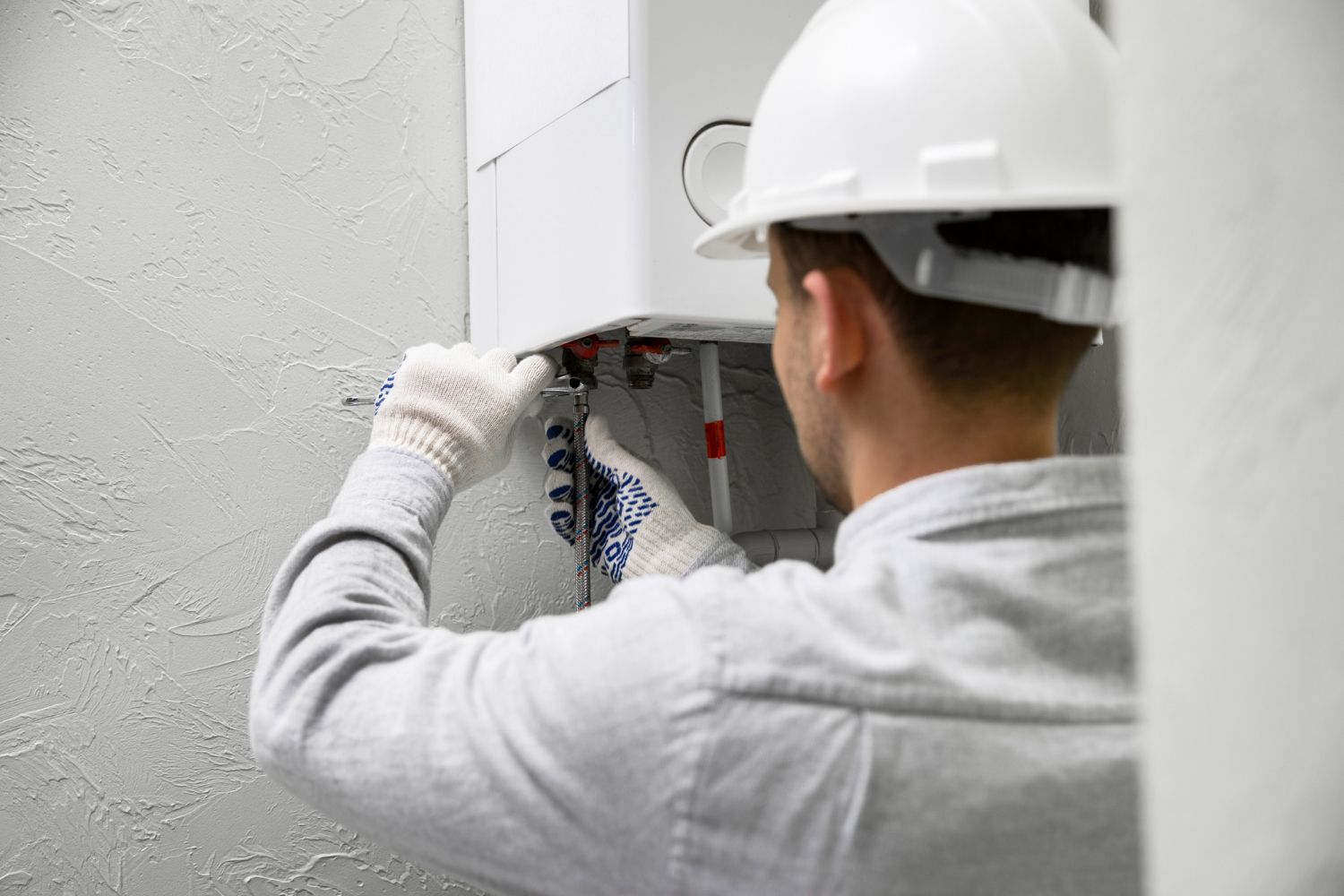 Plumber in a hard hat and gloves working on pipes near a wall-mounted appliance.