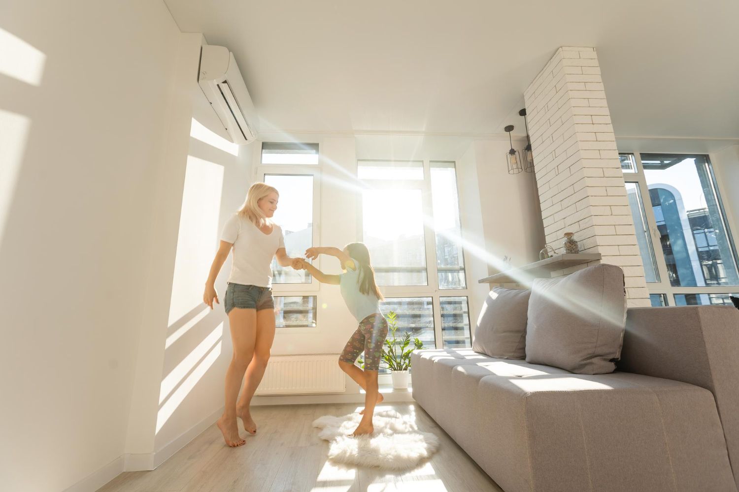 Woman and child dancing in a bright living room, holding hands near window. Sunlight streams in.