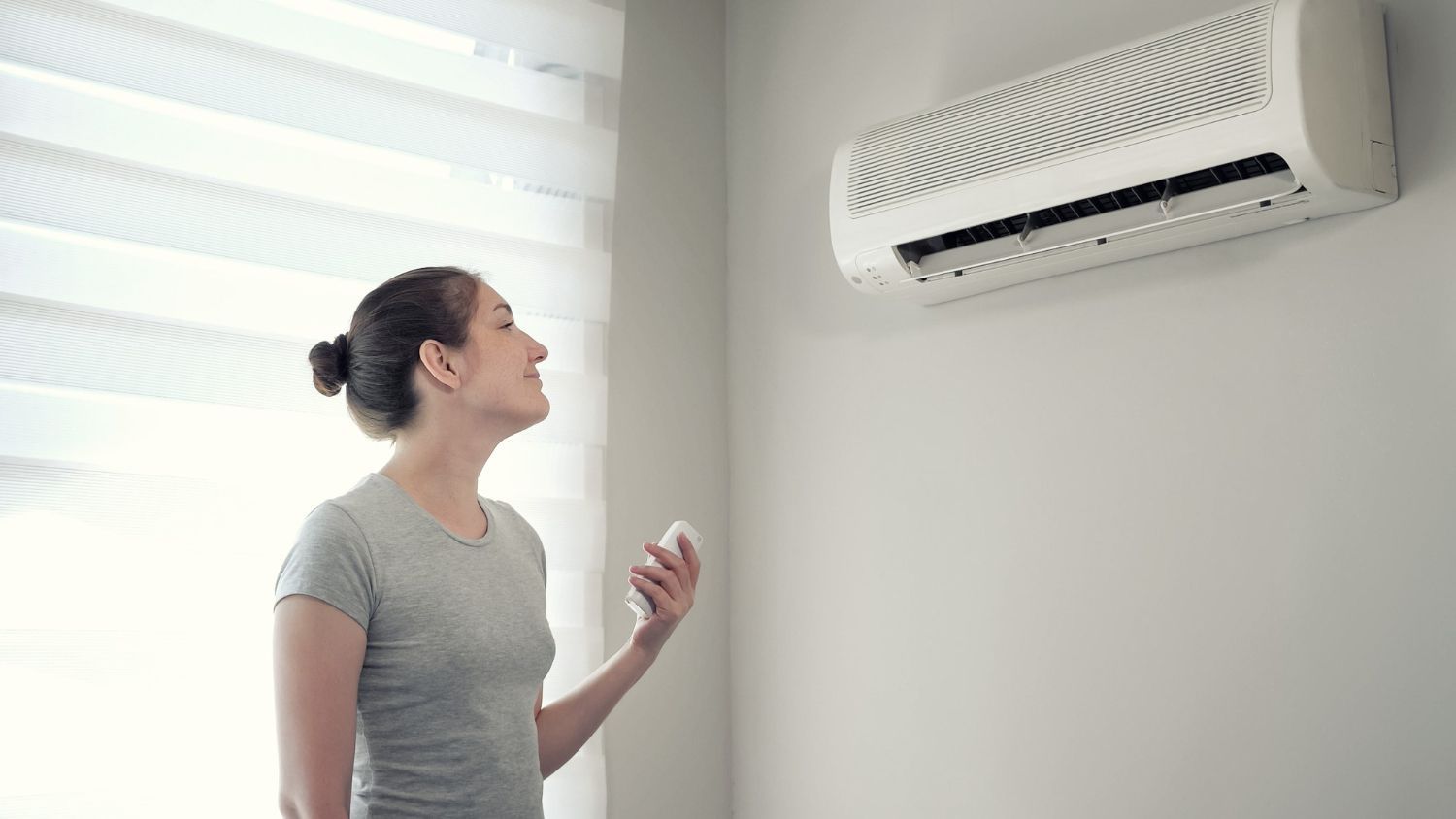 Woman adjusting air conditioner with remote control in a room with a window and white wall.