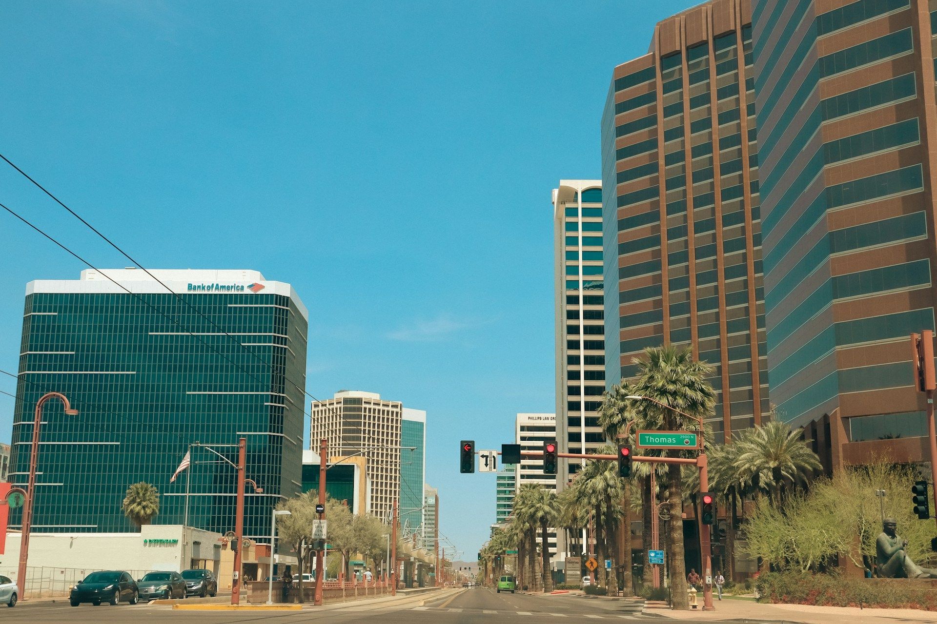 City street view with tall buildings under a clear blue sky. Palm trees and traffic signals are visible.