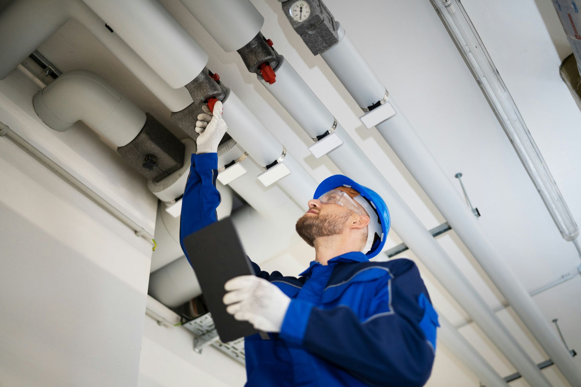 Plumber inspecting pipes overhead, wearing safety gear and holding clipboard.