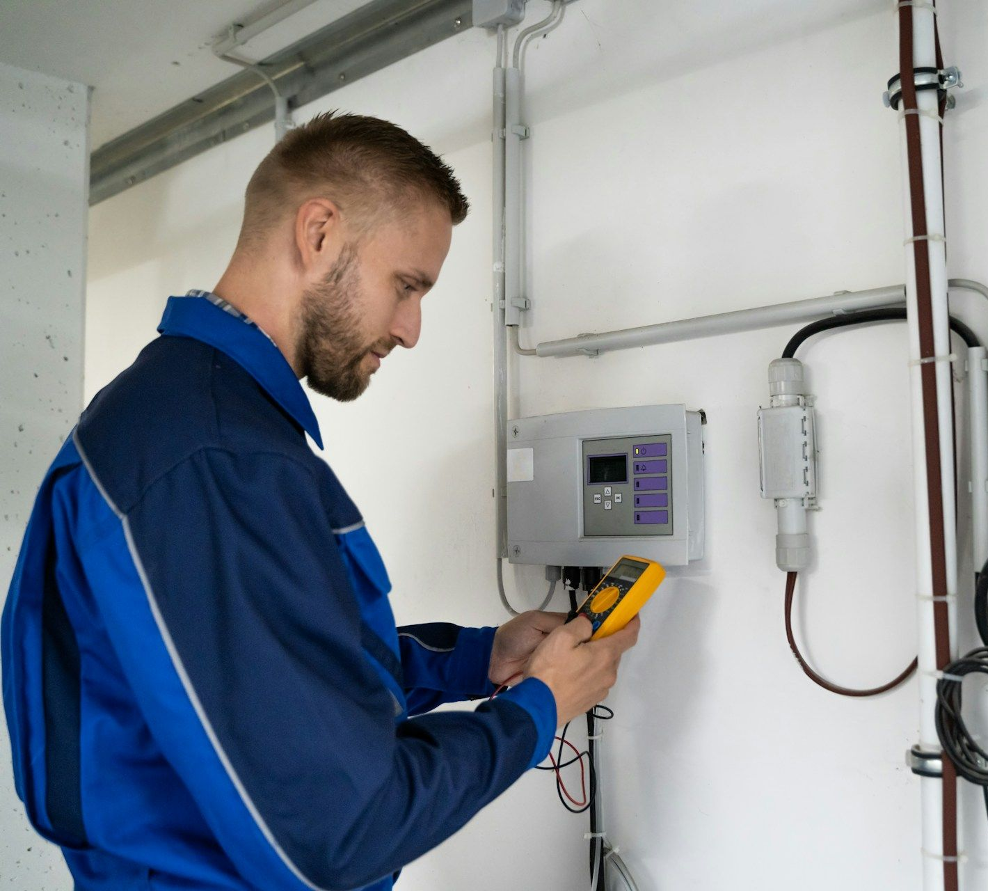 Man in blue overalls using a multimeter on a control panel in an industrial setting.