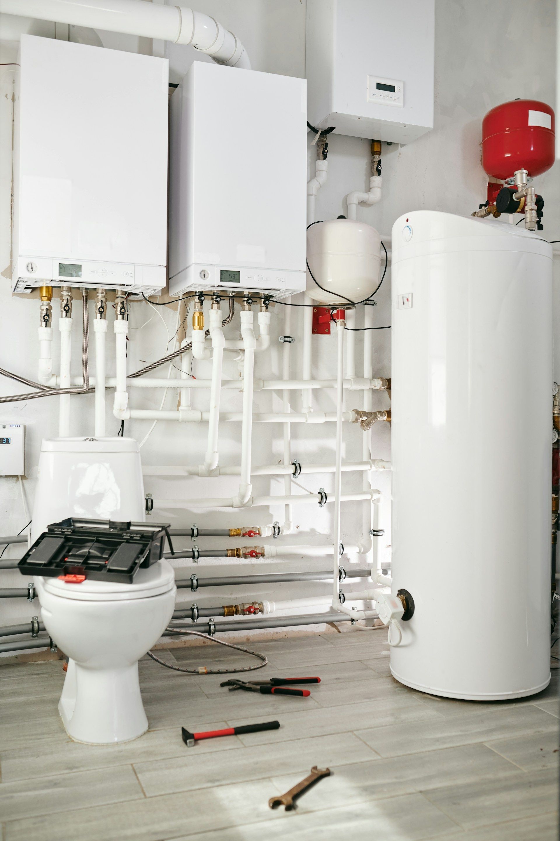 White bathroom with water heaters, pipes, toilet, toolbox, and tools.