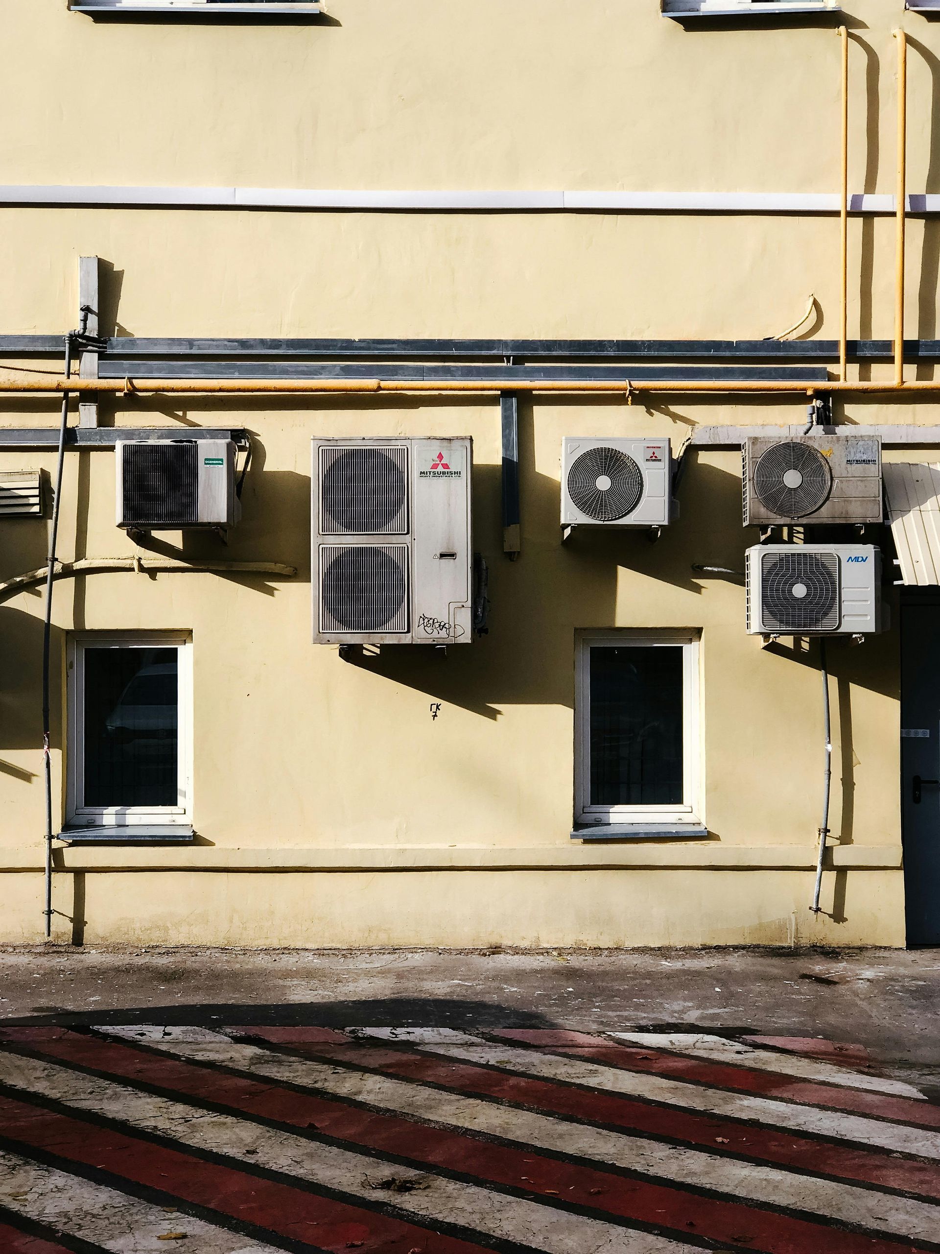 Yellow building wall with several air conditioning units and two windows.