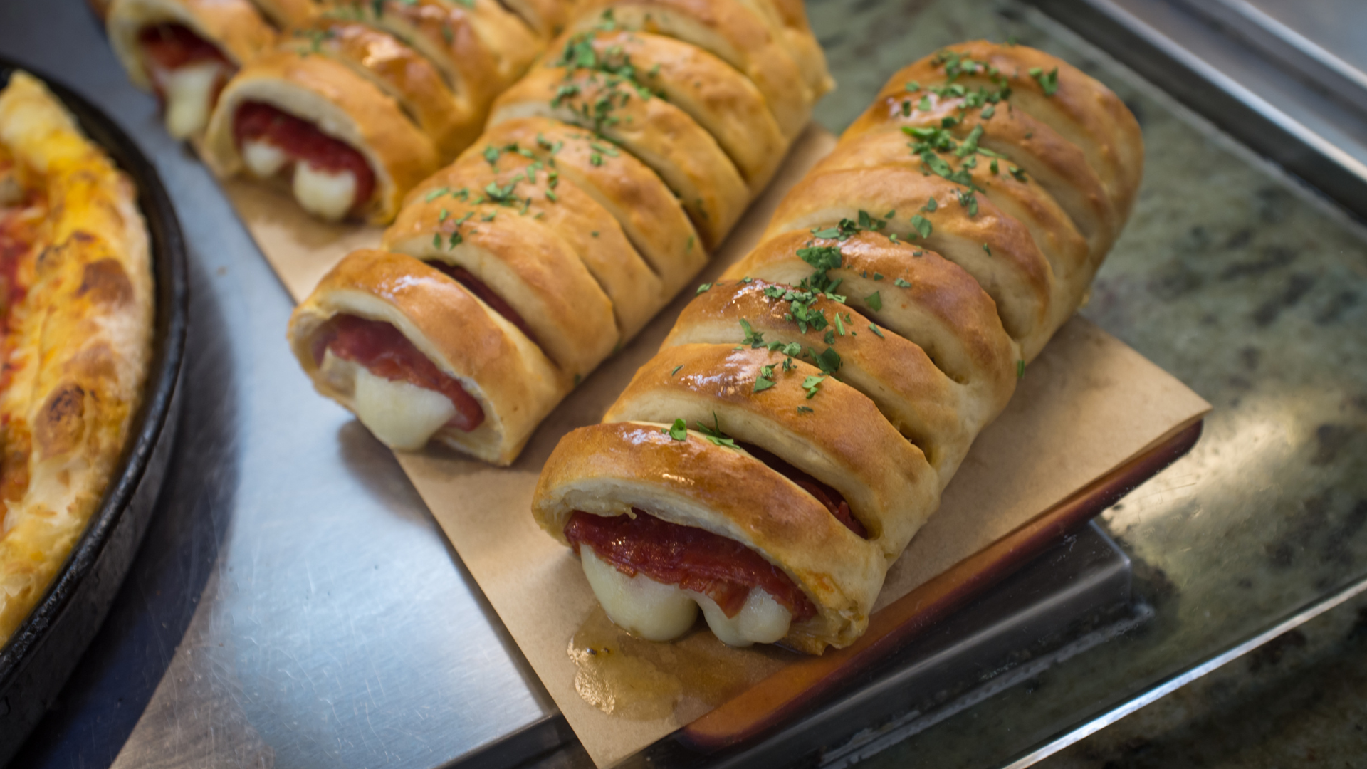 Two baked, sliced strombolis with visible melted cheese and salami, resting on a wooden board next to a pizza.