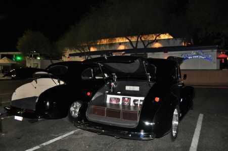 Two classic black cars parked at night with their trunks open, displaying decorative items inside.