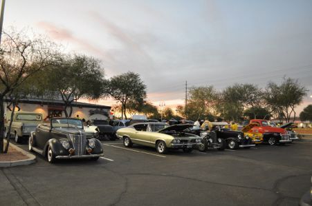 A row of classic cars parked in an outdoor lot at sunset, with people gathering near the vehicles.