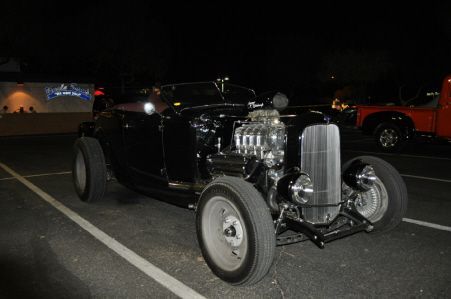A black hot rod with a prominent chrome engine and exposed front wheels parked in a parking lot at night.