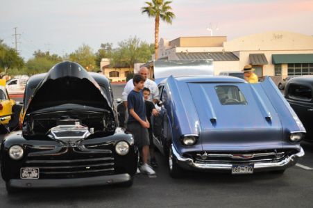 A black car and a metallic blue car, both with their hoods open, parked outdoors with people standing between them.