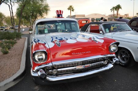 A red and white 1957 Chevrolet with an Uncle Sam-style top hat on the roof, parked at an outdoor car show.