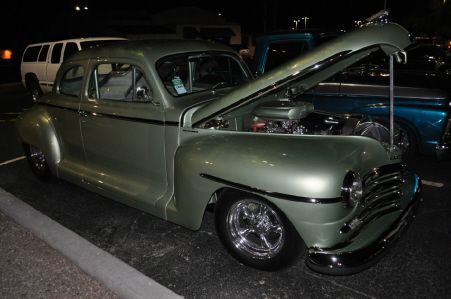 A sage green vintage coupe with a raised hood and chrome wheels parked in a lot at night.