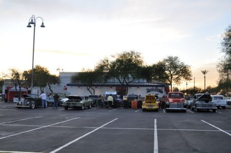 A gathering of classic cars parked in a paved lot in front of a restaurant at sunset.
