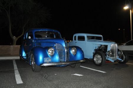 A dark blue vintage car parked next to a light blue hot rod in a parking lot at night.