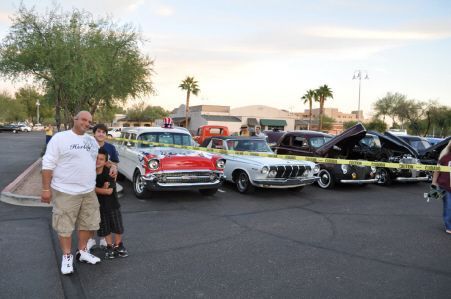 A man and two children pose by a row of classic cars in a parking lot during a car show under a bright, clear sky.
